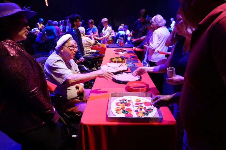 In the Come to the Edge Cafe at the Performing Arts Centre in St. Catharines, John from March of Dimes Brain Injury Unit -a man wearing a white shirt and white chef's cap sitting in wheelchair - speaks to audience member wearing blue shirt as she picks out her fabric pizza from table covered in fabric food
